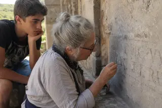 Elderly artisan restoring a limestone inscription in a shaded Puéchabon courtyard, with a local teenager observing, bathed in soft golden light