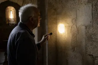 An elderly man in work clothes quietly shining a flashlight on an ancient carving inside a dimly lit Romanesque church, with soft golden light illuminating stone textures and inscriptions.