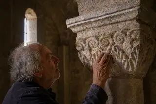 An elderly stonemason studies stone details inside a Romanesque church, gentle light highlighting ancient carvings and his focused expression, creating an intimate, peaceful mood.