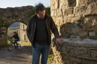 Historian touching ancient stone tower base in quiet village, with child cycling past old ramparts in early morning light.