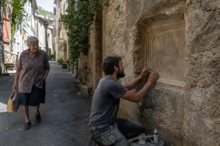 Artisan restoring an inscription on a stone wall in a sunlit alley of Puéchabon, with an elderly resident walking by, both absorbed in their activities.
