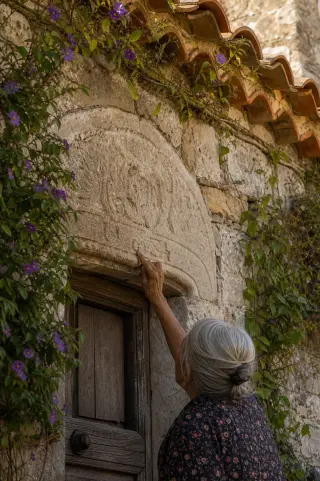Elderly woman examines stone carving on an old decorated doorway in Puéchabon, surrounded by climbing flowers and sunlit textured masonry.