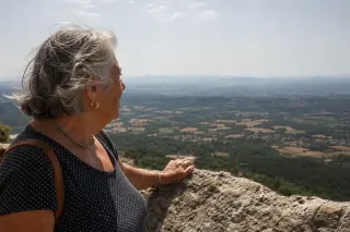 Elderly local woman in contemplation, hand on stone parapet, overlooking panoramic countryside from Rocher de la Vierge under soft Languedoc light