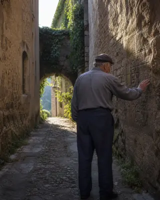 Elderly man gently touching old inscribed stone wall in a narrow medieval alley bathed in soft morning light, with an overgrown archway in the background.