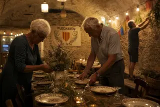 Elderly villagers arranging wildflowers and ceramics on a long table in a stone community hall, with warm lanterns and rustic decorations creating a peaceful, authentic atmosphere.