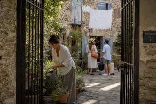 Open courtyard of a historic stone house in Puéchabon, a local woman watering plants as travelers arrive, subtle morning light and calm atmosphere.