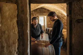 Winemaker and visitor in a stone cellar, absorbed in conversation, with golden light illuminating ancient wine barrels and textured walls.
