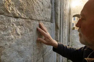 Elderly artisan's hand tracing weathered limestone wall, soft golden light in a stone alley of Puéchabon, conveying authentic connection with the historic architecture.